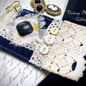 Puede incluir: Una pieza textil artesanal con mezclilla, encaje y un bot&oacute;n negro. El dise&ntilde;o incorpora un imperdible dorado, botones vintage y el texto "Vintage Mama's Cottage". La pieza muestra una mezcla de texturas y materiales.