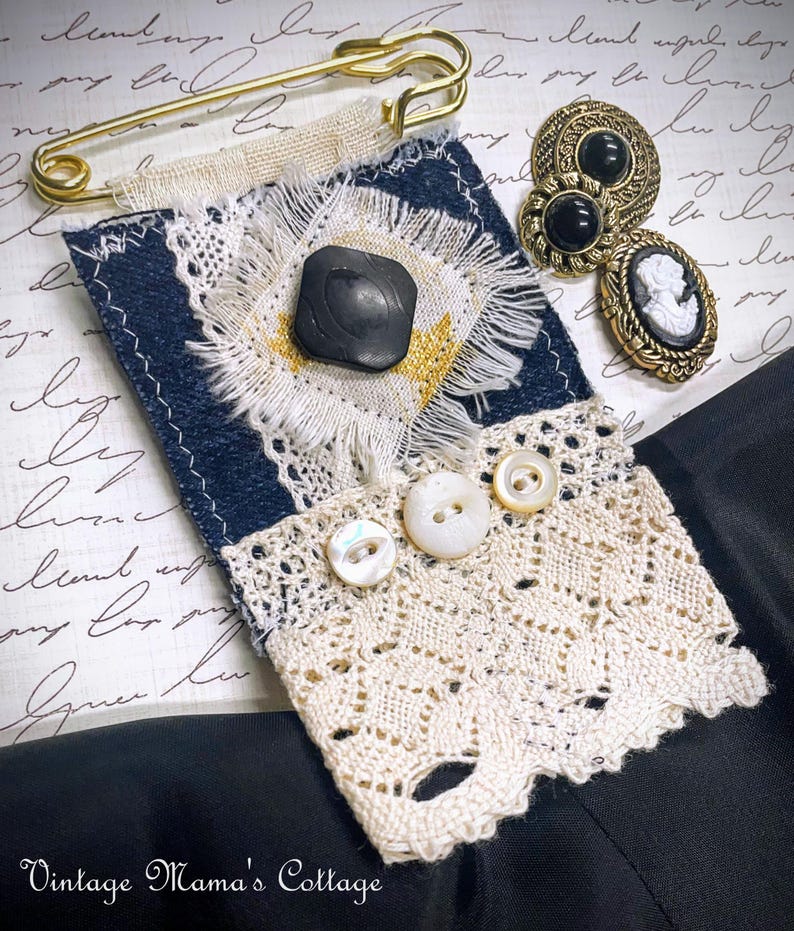 Puede incluir: Una colecci&oacute;n de accesorios de estilo vintage, que incluye un broche de tela azul marino y crema con un bot&oacute;n negro, ribete de encaje y botones de perlas. Tambi&eacute;n hay un imperdible dorado y botones antiguos con piedras negras y un camafeo. El texto "Vintage Mama's Cottage" es visible.