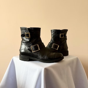 May include: A pair of black leather ankle boots with gold-colored square buckles. The boots have a worn, textured appearance and are displayed on a white surface against a neutral background. The boots have a low heel.