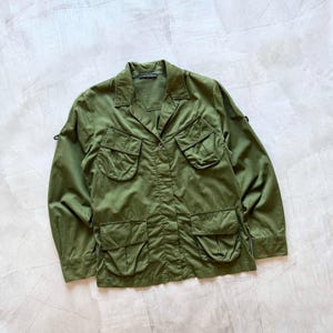 Puede incluir: Chaqueta verde oliva con múltiples bolsillos delanteros y cuello. La chaqueta tiene cierre de botones y mangas largas. La tela parece ligera. El diseño recuerda a una prenda de estilo militar. La chaqueta mide unos 70 cm de largo.