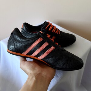May include: A pair of black low-top trainers featuring a perforated leather upper, three bright orange side stripes, matching orange laces, and a thin orange sole. The Adidas logo is visible on the heel tab of the black leather footwear.