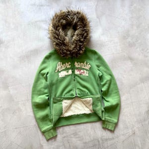 May include: A green Abercrombie zip-up hoodie with a faux fur-lined hood. The hoodie features the brand's logo in white and red lettering. The interior is lined with a cream-coloured, fuzzy material.