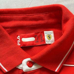 Pu&ograve; includere: Primo piano di una polo rossa con colletto bianco e bottone rosso. La camicia ha una striscia bianca sul colletto e il logo Fila. L'etichetta recita "Scuderia Ferrari" e "Fornitore Ufficiale".