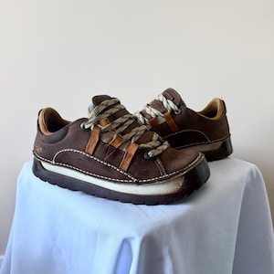 May include: A pair of dark brown leather low-top sneakers featuring a thick, white, textured rubber sole, tan leather accents along the lace eyelets, and beige and brown patterned laces. The shoes have visible white contrast stitching.