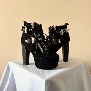 May include: A pair of black ankle boots with high block heels and thick platforms. The boots feature silver-toned metal buckles, side zippers, and black laces threaded through metal eyelets. The exterior is a matte black material with a textured finish.