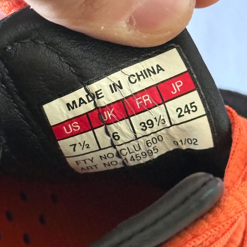 May include: A white rectangular shoe size label with black text reading MADE IN CHINA, US 7 1/2, UK 6, FR 39 1/3, JP 245, FTY NO. CLU 600, ART NO. 145995, and 01/02. The label is attached to the interior of a black and orange athletic shoe.