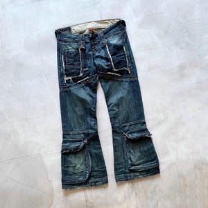 May include: Dark blue denim jeans with distinctive pocket and seam detailing. The jeans have multiple pockets, decorative stitching, and a button-fly. The design includes distressed elements and a flared leg opening, offering a vintage aesthetic.