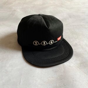 Könnte beinhalten: Schwarze Baseballkappe mit flachem Schirm. Auf der Kappe sind die Zahlen 2, 3 und 4 in weißen Kreisen abgebildet, gefolgt von Auslassungspunkten. Ein kleines rot-weißes Logo befindet sich an der Seite.
