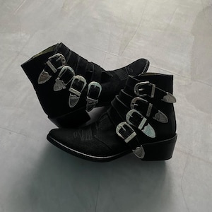 Op de afbeelding: Een paar zwarte suède enkellaarsjes met zilveren gesps. De laarsjes hebben een spitse neus en een lage hak.