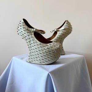 May include: A pair of off-white platform shoes featuring a high, curved heel and a Mary Jane strap with a metal buckle. The entire exterior surface of the shoes is densely covered in small, sharp, silver-toned metal spikes and studs.