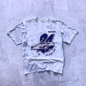 Könnte beinhalten: Weißes T-Shirt mit einem verblassten Tie-Dye-Muster. Das Shirt zeigt eine lila und goldene Grafik eines Rennwagens mit der Nummer 24 und dem Text "24 Heures du Mans" und "La Leggenda".