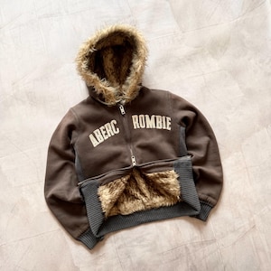 May include: A brown zip-up hoodie with a faux fur-lined hood and pocket trim. The hoodie features the text "ABERC ROMBIE" in white block letters across the chest. The cuffs and bottom hem are a darker grey colour.