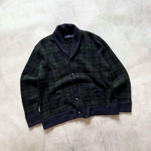 May include: A dark green and navy blue plaid cardigan sweater with a shawl collar and button-up front. The sweater has two front pockets and navy blue trim. The sweater is made of a soft, comfortable material.