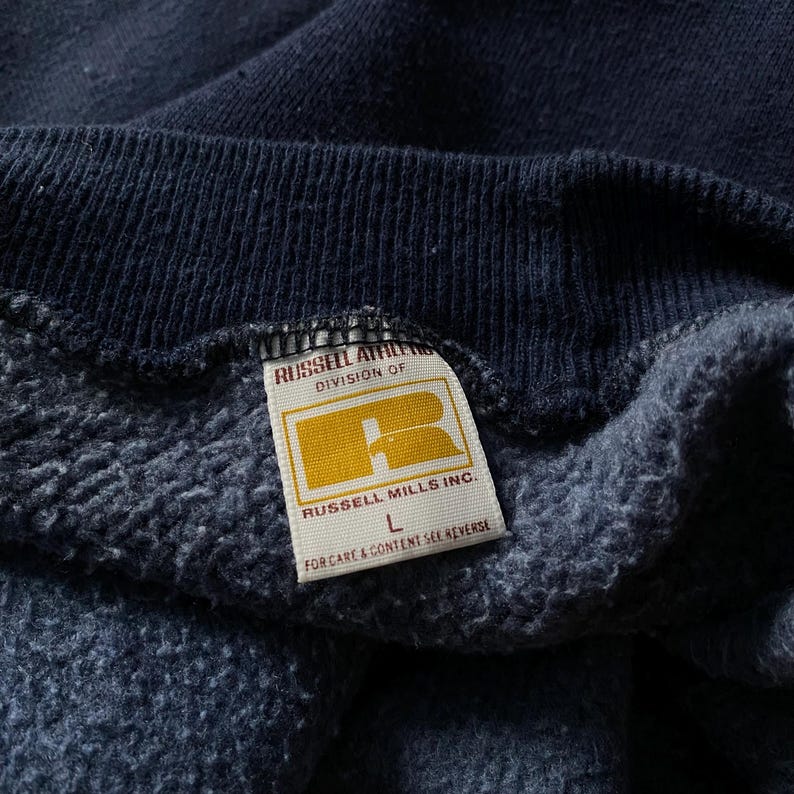 May include: A navy blue sweatshirt with a white tag that reads "Russell Athletic, Division of Russell Mills Inc., L, For care & content see reverse."