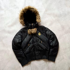 Peut inclure: Veste de bombardier noire avec capuche doublée de fausse fourrure. La veste présente un design matelassé, un ourlet côtelé et des fermetures éclair et boutons dorés. La veste porte les lettres dorées "Ru" brodées sur la poitrine.