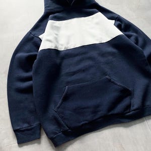 May include: A navy blue hooded sweatshirt with a white horizontal stripe across the chest. The sweatshirt has a kangaroo pocket and long sleeves.