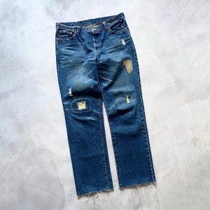 Op de afbeelding: Blauwe denim jeans met distressed details, waaronder scheuren en patches. De jeans hebben een klassiek ontwerp met vijf zakken, een knoop- en ritssluiting en een rauwe zoom. De jeans liggen plat op een lichtgrijze ondergrond.