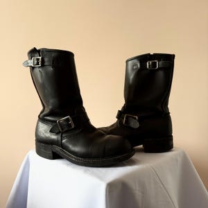 Puede incluir: Un par de botas de moto de cuero negro con hebillas plateadas. Las botas son de media caña y tienen la punta redondeada. Se exhiben sobre una superficie blanca sobre un fondo neutro.