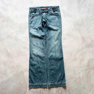 Puede incluir: Vaqueros de mezclilla azul descolorido con cintura baja y pernera acampanada. Los vaqueros presentan dos bolsillos delanteros, detalles de anillos metálicos y detalles de costuras verticales en la parte delantera y trasera de las perneras. Un pequeño logotipo rojo es visible en la parte delantera.