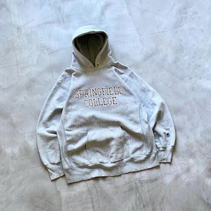 Puede incluir: Sudadera con capucha gris claro con el texto "SPRINGFIELD COLLEGE" en letras granates. Tiene un bolsillo delantero y un logotipo de Champion en la manga izquierda. La capucha está forrada con una tela gris más clara.