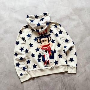 May include: Cream-coloured hoodie with a pattern of navy blue stars. The front features a cartoon monkey dressed as Uncle Sam, wearing a top hat and bow tie. The hoodie has a hood with a star pattern.