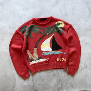 May include: Red knit sweater featuring a sailboat and palm tree design. The sweater has a crew neck and long sleeves. The sailboat has a black sail, and the palm trees are green. The text "RL 20" is at the bottom.
