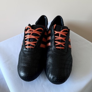 May include: A pair of black athletic trainers featuring a perforated leather upper, three side stripes in a vibrant coral orange, and matching coral orange laces. The low-top shoes have a rounded toe design and a structured heel collar.