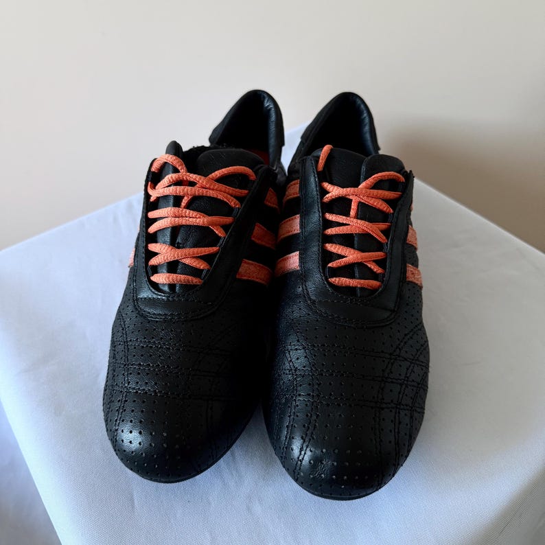 May include: A pair of black athletic trainers featuring a perforated leather upper, three side stripes in a vibrant coral orange, and matching coral orange laces. The low-top shoes have a rounded toe design and a structured heel collar.