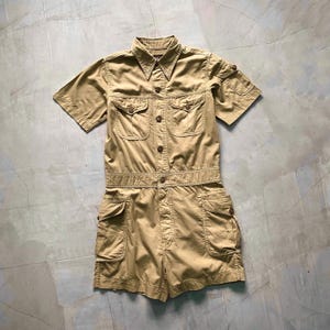 Puede incluir: Un mono utilitario de manga corta de color beige con múltiples bolsillos y cierre de botones en la parte delantera.