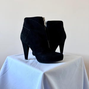 May include: A pair of black suede ankle boots featuring a high stiletto heel, a rounded toe, and a prominent silver-toned metal zipper running vertically down the center front of each boot. The design includes a structured collar and smooth finish.