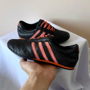 May include: A pair of black athletic trainers featuring a perforated leather upper, bright orange laces, and three prominent orange stripes along the side. The low-top shoes have a slim profile with a thin orange sole and an orange logo on the heel.