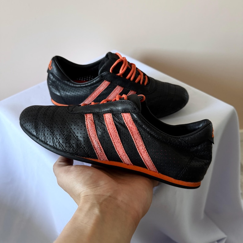 May include: A pair of black athletic trainers featuring a perforated leather upper, bright orange laces, and three prominent orange stripes along the side. The low-top shoes have a slim profile with a thin orange sole and an orange logo on the heel.