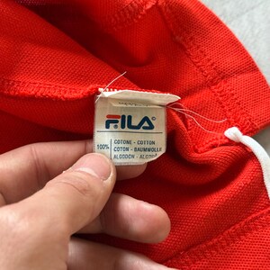 Pu&ograve; includere: Primo piano di un capo rosso con coulisse bianca. Un'etichetta bianca mostra il logo FILA e il testo "COTTON" in pi&ugrave; lingue. Il capo &egrave; realizzato in 100% cotone.