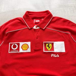 Pu&ograve; includere: Polo rosso Fila con dettagli bianchi e colletto. Presenta loghi ricamati, tra cui Vodafone, Shell e l'emblema Ferrari. La maglia ha una chiusura a bottoni e il logo Fila.