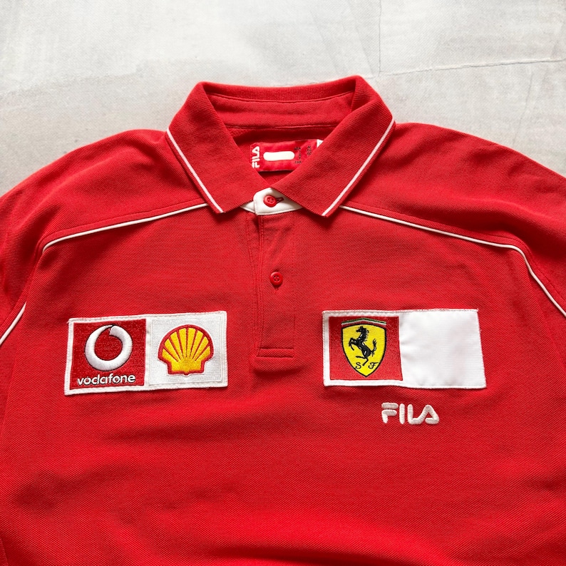 Pu&ograve; includere: Polo rosso Fila con dettagli bianchi e colletto. Presenta loghi ricamati, tra cui Vodafone, Shell e l'emblema Ferrari. La maglia ha una chiusura a bottoni e il logo Fila.