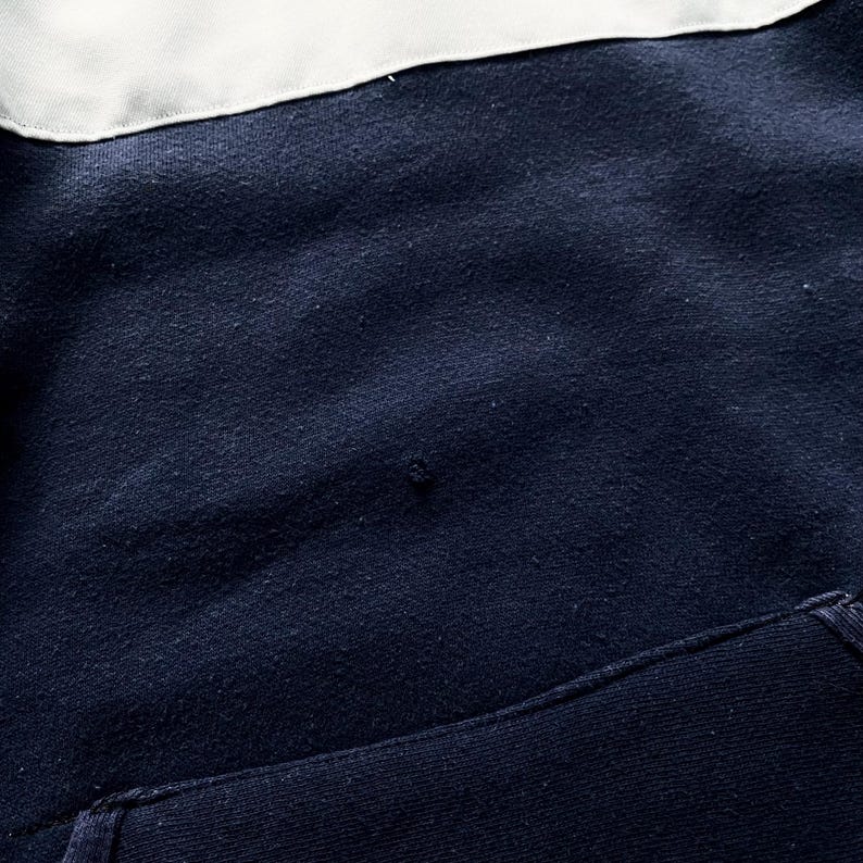 May include: Close-up of a navy blue fabric with a white fabric panel at the top. The fabric has a textured, slightly rough surface.