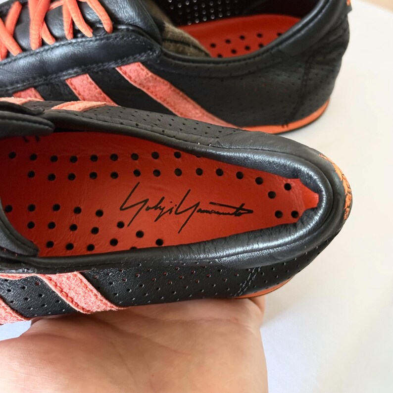 May include: A black leather trainer with perforated detailing and bright orange accents. The interior insole is orange with black perforations and features the signature Yohji Yamamoto printed in black script on the heel area of the shoe.