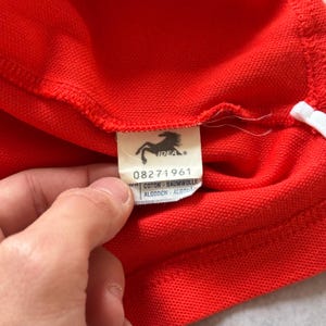 Pu&ograve; includere: Primo piano di una polo rossa con un dettaglio a righe bianche. L'etichetta della maglia presenta un logo a forma di cavallo e la parola "IDEA", insieme al numero "08271961" e testo in pi&ugrave; lingue.