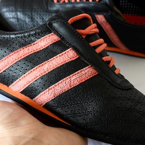 May include: A black perforated leather trainer featuring three textured coral-orange stripes on the side, matching coral-orange laces, and a coral-orange sole. The side of the shoe displays a signature in silver script lettering.