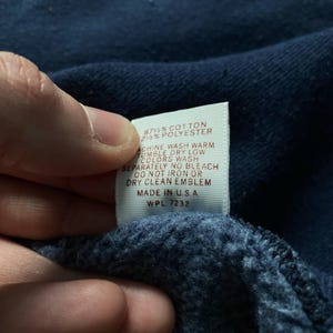 May include: A white tag with brown text detailing the fabric content and care instructions for a garment. The tag reads "87% Cotton 2% Polyester", "Machine Wash Warm", "Tumble Dry Low", "Colors Wash Separately", "No Bleach", "Do Not Iron Or Dry Clean Emblem", "Made in USA", "WPL 7232".