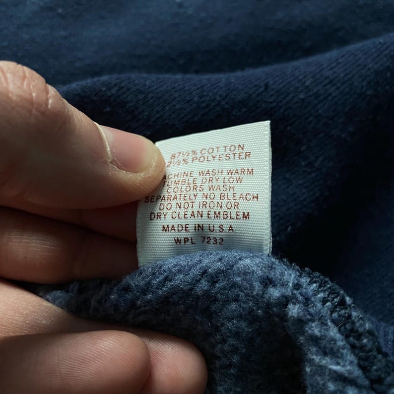 May include: A white tag with brown text detailing the fabric content and care instructions for a garment. The tag reads "87% Cotton 2% Polyester", "Machine Wash Warm", "Tumble Dry Low", "Colors Wash Separately", "No Bleach", "Do Not Iron Or Dry Clean Emblem", "Made in USA", "WPL 7232".