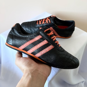 May include: A pair of black low-top trainers featuring a perforated leather upper, three bright orange side stripes, matching orange laces, and a thin orange sole. The design includes a rounded toe and a padded collar for ankle support.