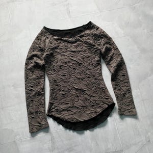 May include: A long-sleeved, brown top with a unique, textured design featuring numerous small, eye-like cutouts. The top has a scoop neck and a black lining visible at the neckline and hem. The fabric appears to be soft and draped.