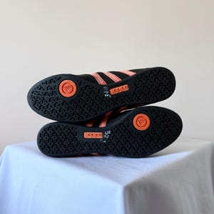 May include: A pair of black athletic shoes with coral-coloured stripes and soles. The black rubber outsoles feature a textured pattern, a circular coral insert with the Adidas logo, and a rectangular coral panel with the Adidas brand name.
