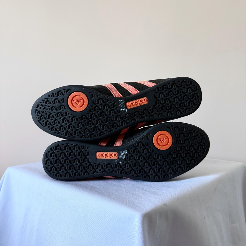 May include: A pair of black athletic shoes with coral-coloured stripes and soles. The black rubber outsoles feature a textured pattern, a circular coral insert with the Adidas logo, and a rectangular coral panel with the Adidas brand name.