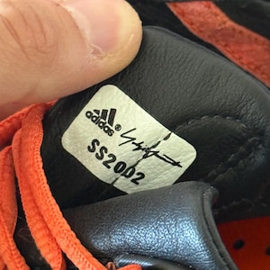 May include: A black leather shoe interior featuring a white rectangular label with the Adidas logo, a signature, and the text SS2002 printed in black. Bright orange laces are visible along the side of the black textured leather material.