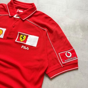 Pu&ograve; includere: Polo rosso con dettagli bianchi e loghi ricamati. Presenta il logo Ferrari, Shell e Vodafone. La maglietta ha un colletto e maniche corte.