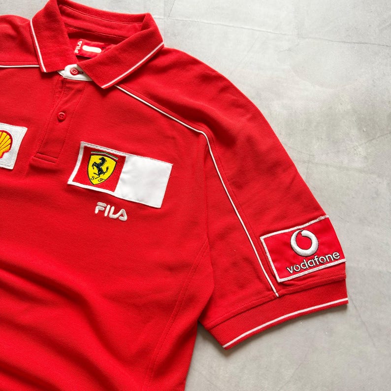 Pu&ograve; includere: Polo rosso con dettagli bianchi e loghi ricamati. Presenta il logo Ferrari, Shell e Vodafone. La maglietta ha un colletto e maniche corte.