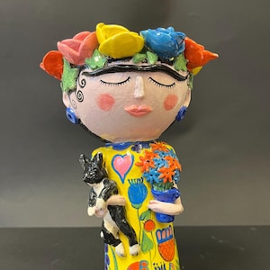 Puede incluir: Una figurita de cerámica de una mujer con un vestido amarillo y una corona de flores. Lleva un gato blanco y negro y una maceta azul con flores.