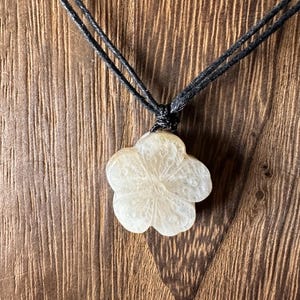 May include: A cream-colored flower pendant necklace with a black cord. The flower pendant has a detailed carved design. The necklace is displayed against a brown wooden background.